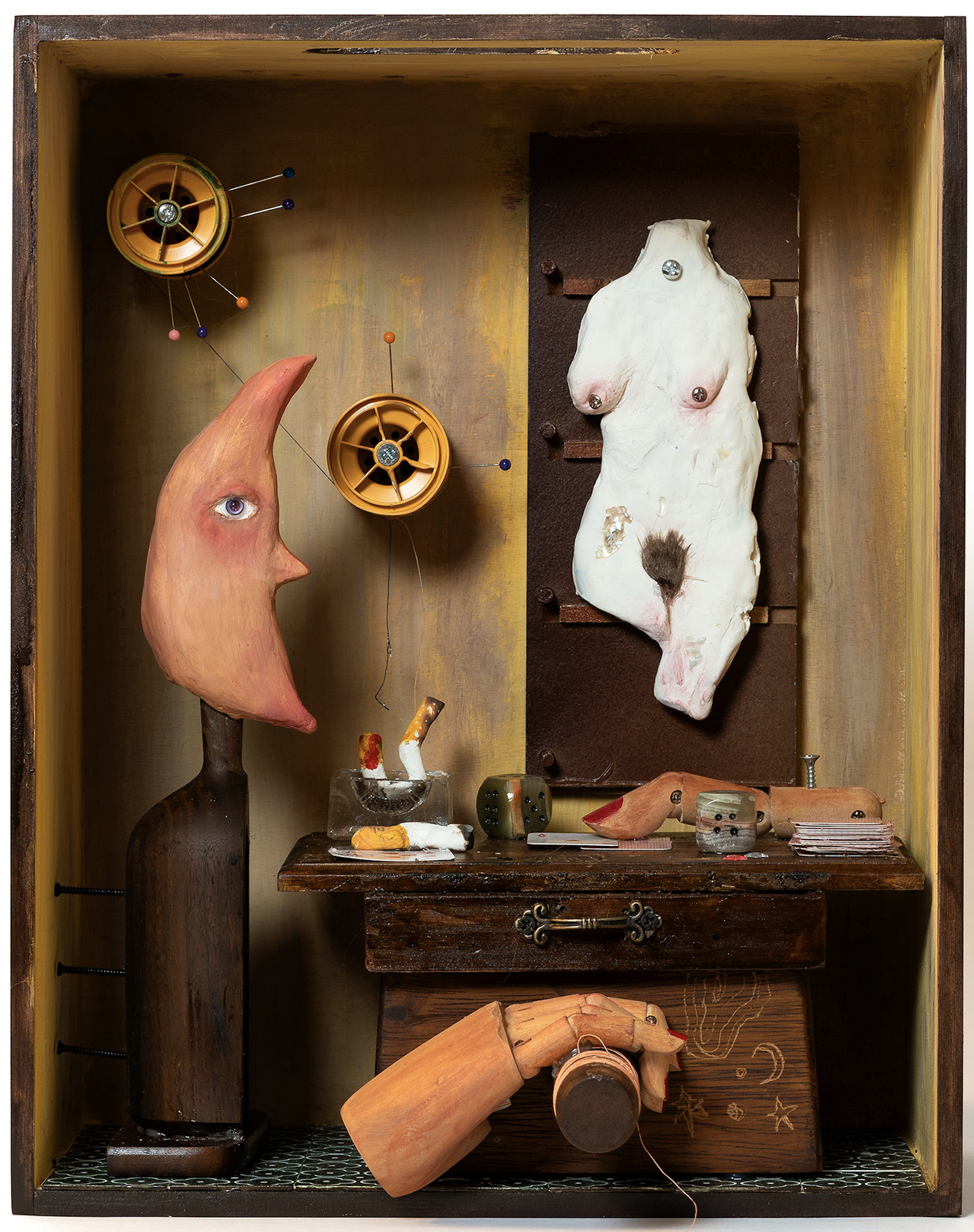 Shadow box sculpture featuring a crescent moon face, a sculpted torso, a tabletop with miniature cards, dice, and a smoking cigarette. A wooden mannequin hand reaches out, creating a surreal, uncanny scene.